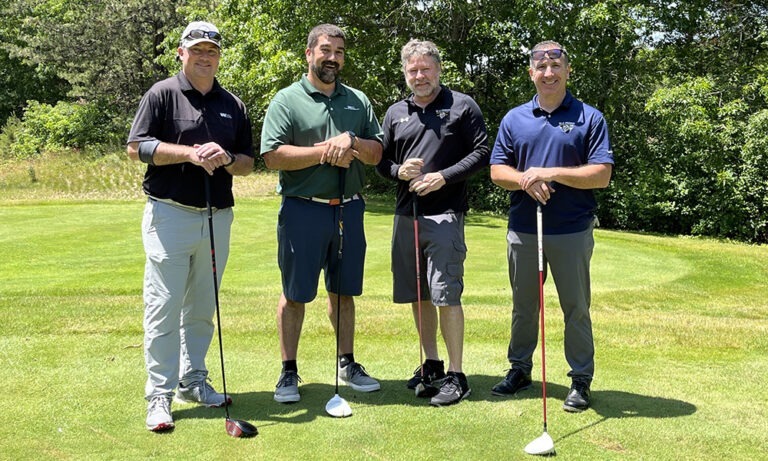 Group of four men on the golf course, holding golf clubs and smiling during the thirteenth annual charity golf tournament.