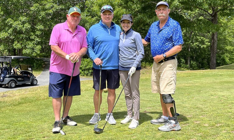 Group of three men and a woman on the golf course, holding golf clubs and smiling during the thirteenth annual charity golf tournament.